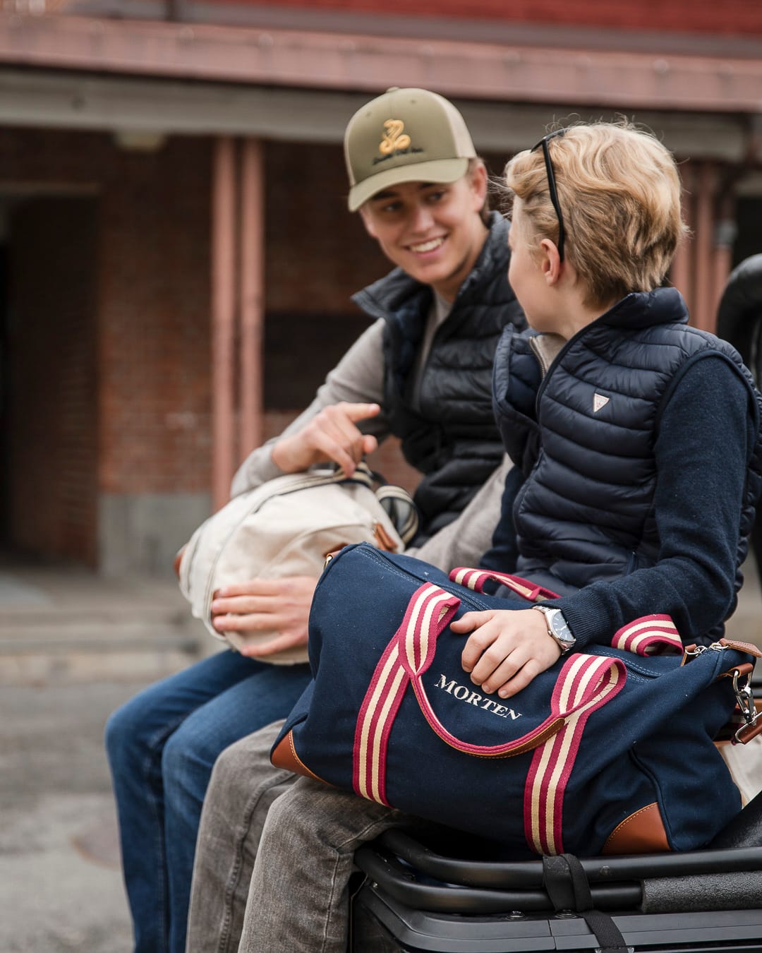To gutter sitter foran en skole. Guttene har hver sin retro-inspirerte bag i fanget. En beige og en blå.