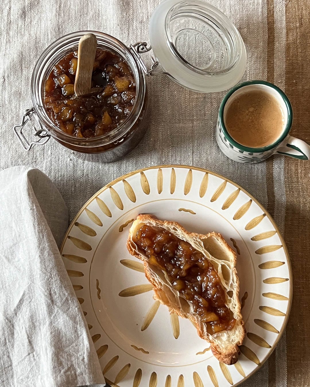 Glass med sykteteøy, kaffekopp og croissant med pæresyltetøy på rustik tallerken sett ovenifra.