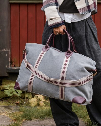 Nærbilde av grå reiseveske med skrå bånd som går over i håndtak i skinn. Båndene er hvite med burgunderrøde striper.