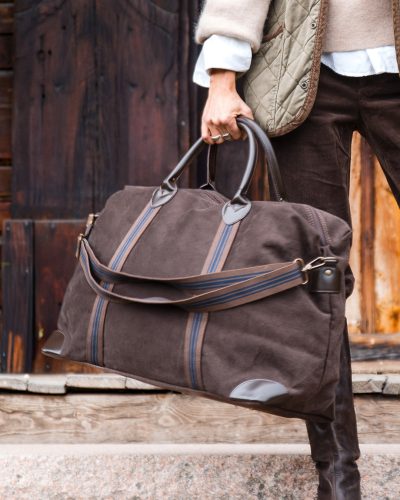 Kvinne holder stor mørkebrun reiseveske i bomullscanvas. Vesken er dekorert med skråstilte bånd med blå striper som møter håndtaket i brunt skinn.