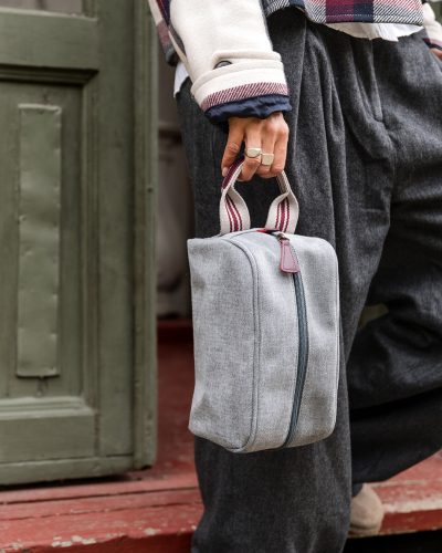 Kvinnehånd holder grå toalettmappe med hvitt håndtak med burgunderrøde detaljer.