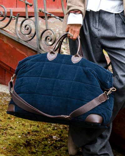 nærbile av reiseveske i mørkeblå cord i et rustikt miljø. Vesken holdes av en kvinnehånd.