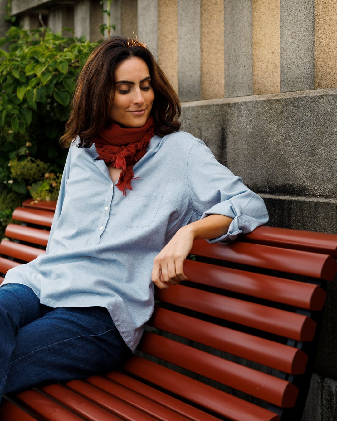 Kvinne med mørkt hår sitter på rød benk og lener armen over ryggstøtten. Hun har på seg en lyseblå skjorte i bomullsflanell og et rødt skjerf i halsen.
