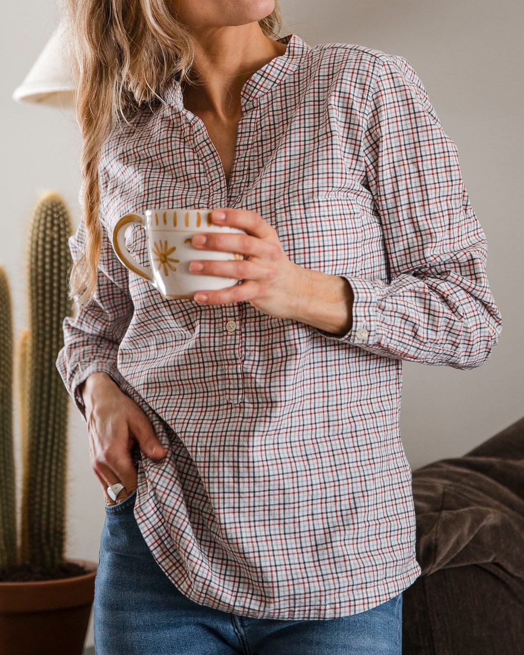 Kvinne med lyst hår holder en tekopp i den ene hånden. Hun har på seg en smårutet skjorte i hvitt, rødt og brunt.