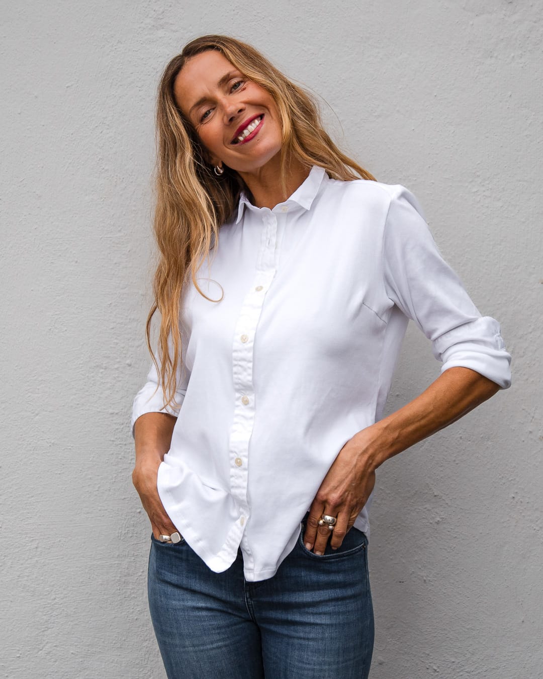 Kvinne med lyst hår har på seg en hvit skjorte i myk pimabomull med knappestolpe, krage og mansjetter i egyptisk bomull og blå jeans. Kvinnen har nedene i lommen og smiler mot kamera.