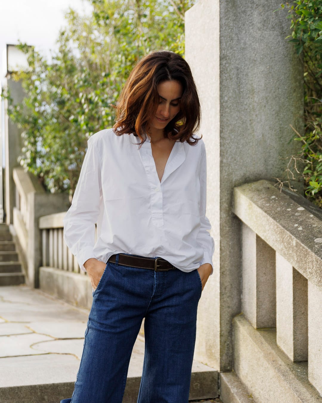 Kvinne med mørkt hår har på seg en oversized Salerno-skjorte i hvitt. Hun står oppreist i en trapp med grønn vegetasjon i bakgrunnen.